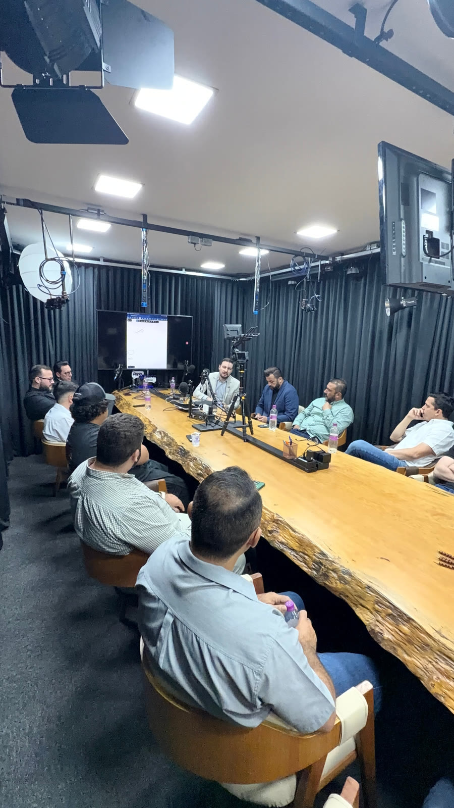 Estúdio com câmera e equipe de luz montadas enquanto um empresário fala à mesa